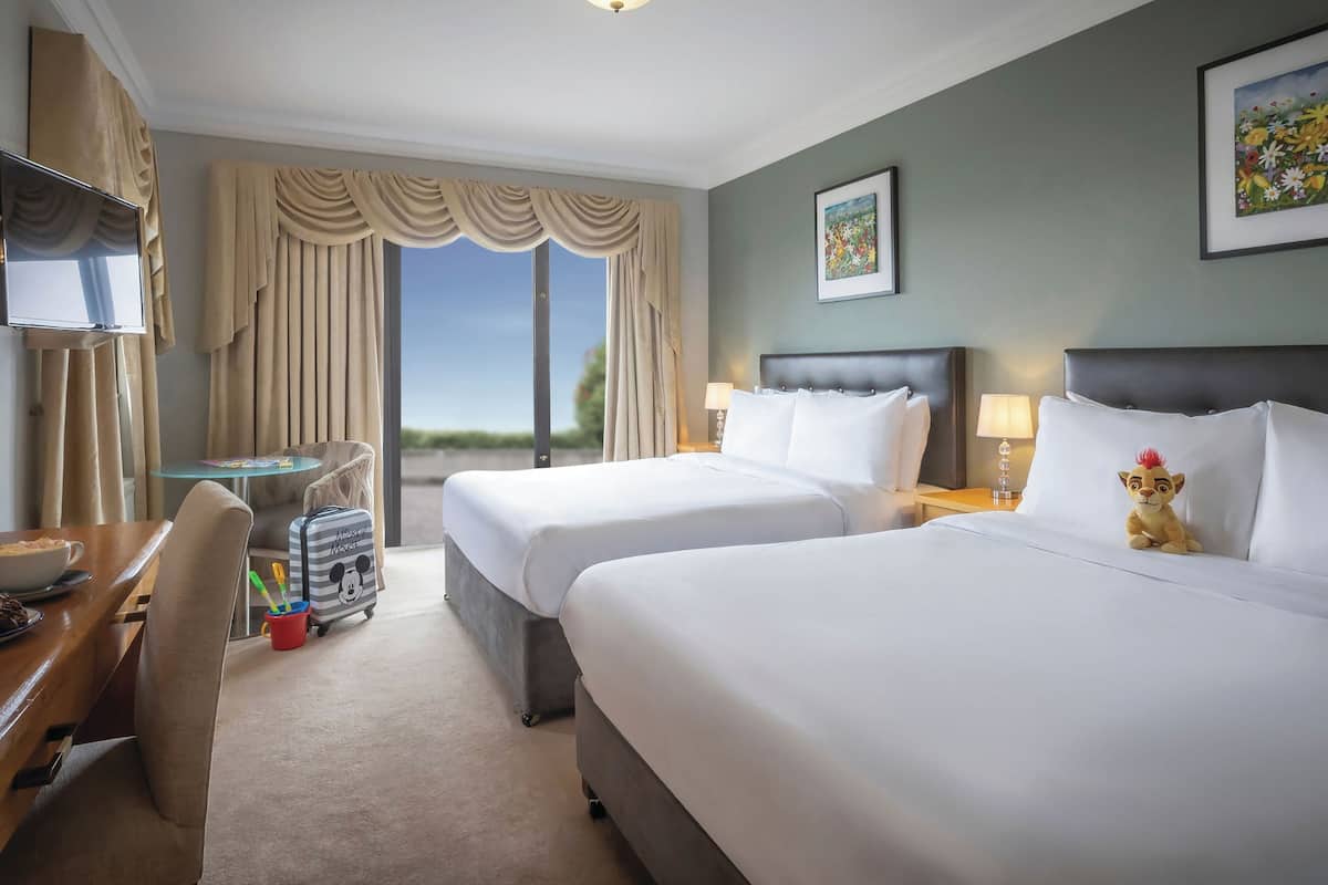 A hotel room with two beds, a desk, a chair, a TV, and a window with curtains.