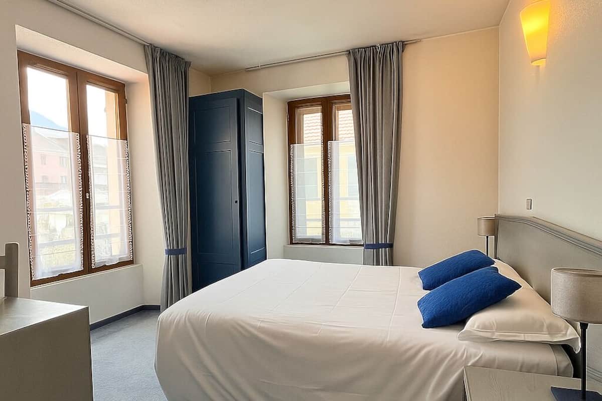 Ein Hotelzimmer mit einem großen Bett, zwei Fenstern mit Vorhängen, einem Kleiderschrank und einer Nachttischlampe.