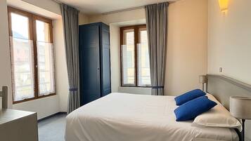 A hotel room with a large bed, two windows with curtains, a wardrobe, and a bedside lamp.
