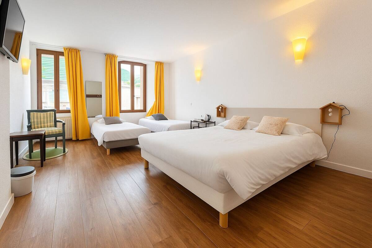 Ein Hotelzimmer mit einem großen Bett, zwei Einzelbetten, einem Sessel, einem Tisch und einem fenster mit Vorhängen.