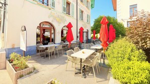 An outdoor seating area with tables and chairs, red umbrellas, and a building with a sign that reads "Hotel De Genevois".