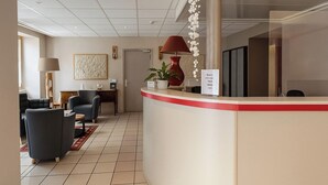 A hotel reception area with a curved reception desk, a red lamp, a vase with white flowers, and a seating area with two armchairs and a small table.