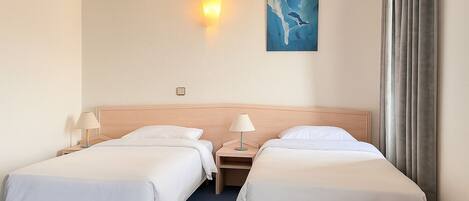 A hotel room with two beds, a nightstand with a lamp, a painting on the wall, and a window with curtains.