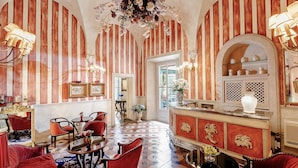 Un hall d’hôtel aux murs rayés, avec des fauteuils à revêtement rouge, un sol à carreaux et un coin bar agrémenté d’un vase blanc et d’objets décoratifs.