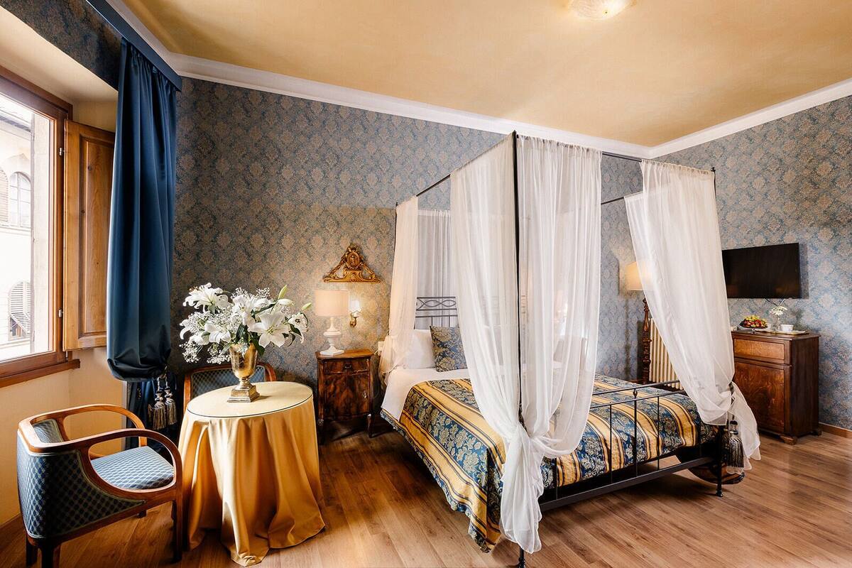 A four-poster bed with a canopy, a round table with a vase of flowers, a chair, and a bedside table with a lamp.