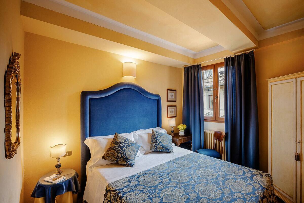 A bedroom with a large bed, a nightstand, a lamp, a window with curtains, and a wardrobe.