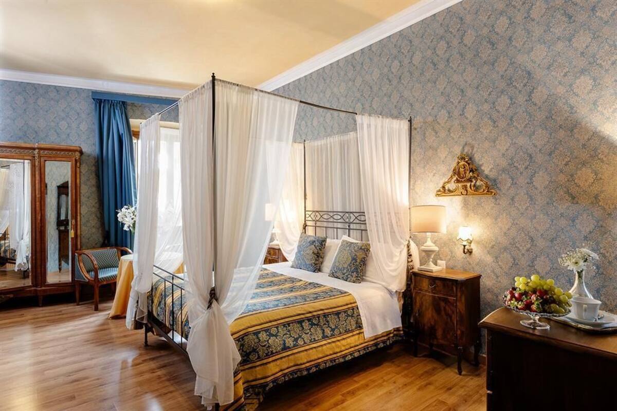 A four-poster bed with white drapes, a wooden nightstand with a lamp, a mirror, and a floral arrangement on a dresser.