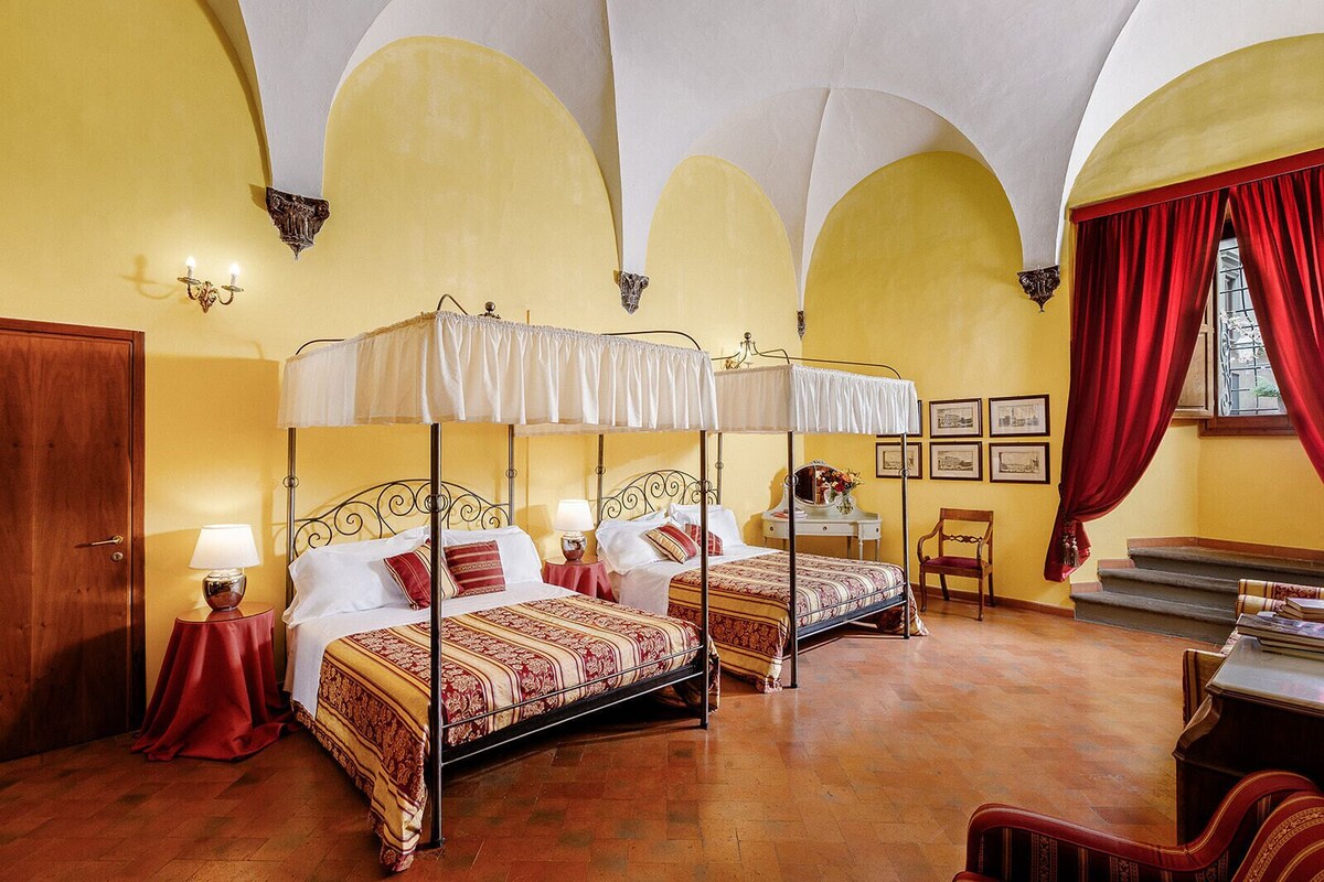 A room with two four-poster beds, a wooden wardrobe, a desk with a chair, and a window with red curtains.