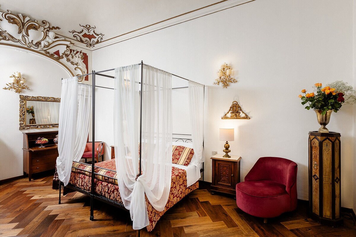 A four-poster bed with a canopy, a red armchair, a wooden cabinet with a vase of flowers, and a wall-mounted lamp.