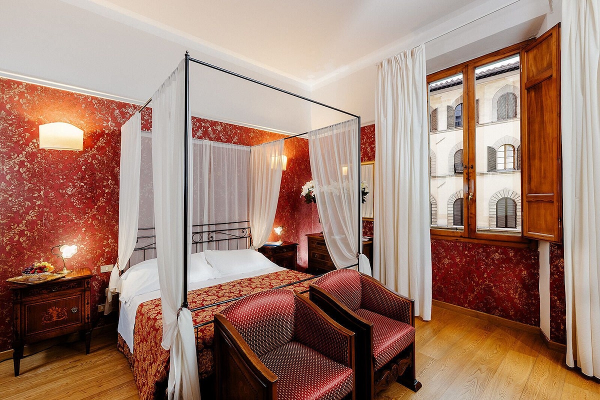 A four-poster canopy bed with a red patterned bedspread, a wooden nightstand with a lamp, and a red upholstered chair.