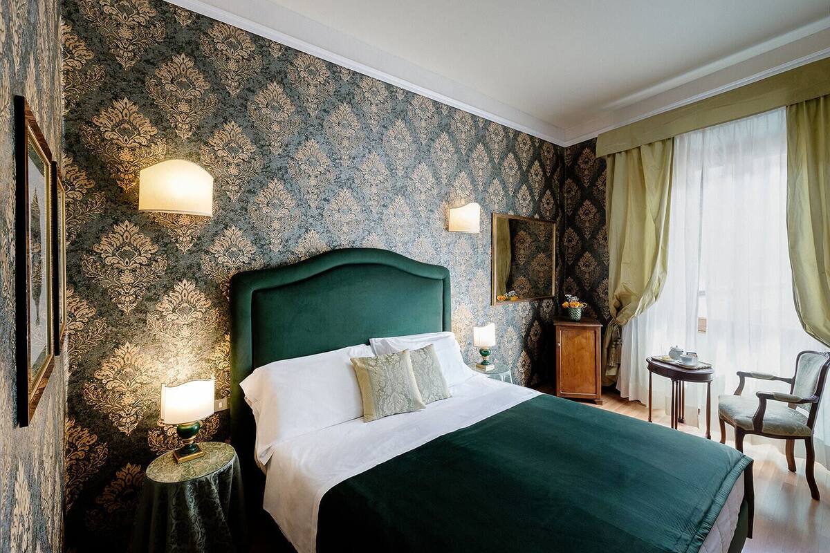 A hotel room with a large bed, two bedside tables, a nightstand, a dresser, a chair, and a window with curtains.