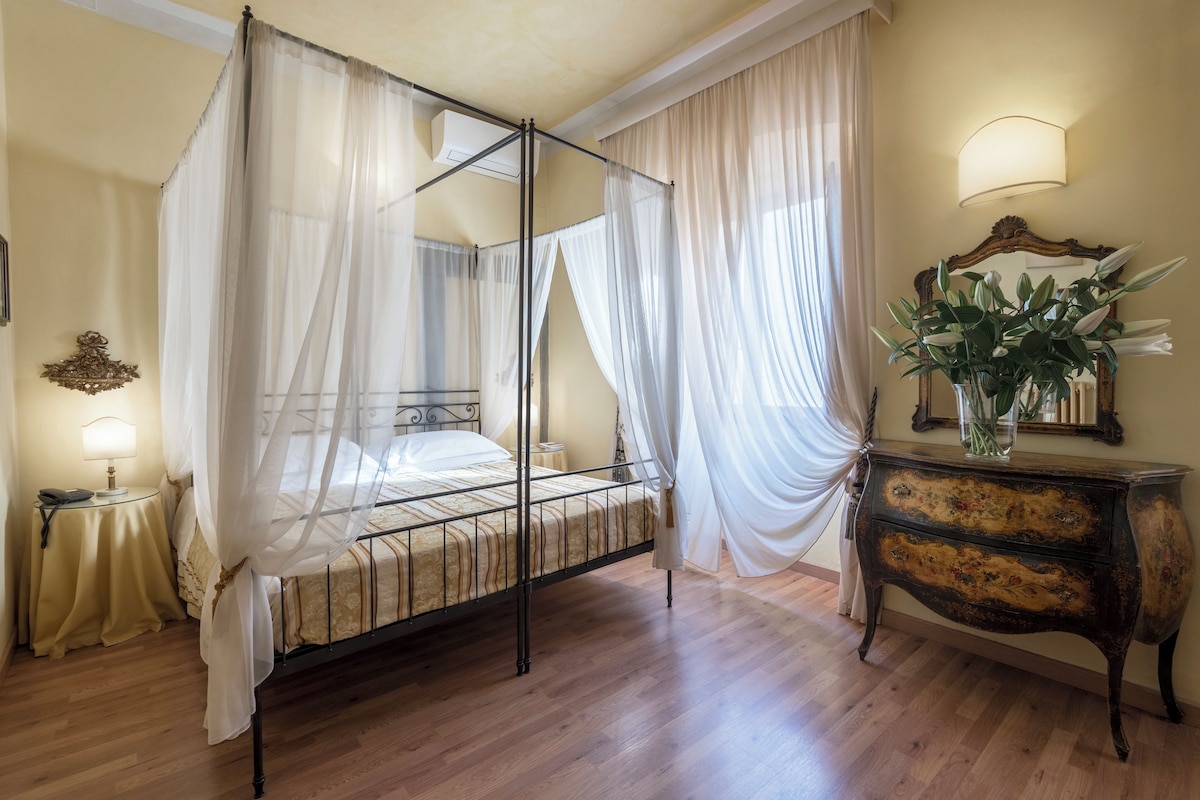 A four-poster bed with a canopy, a bedside table with a lamp, a wooden dresser, and a mirror on the wall.