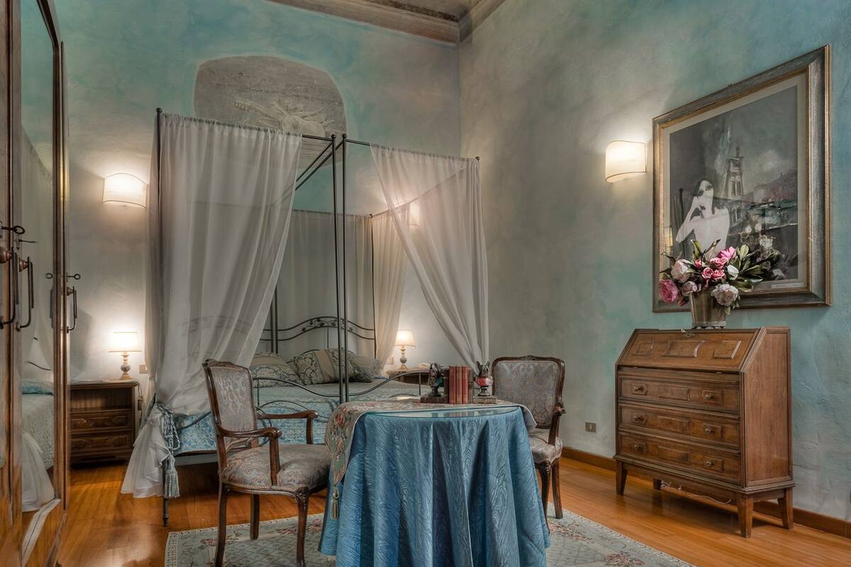 A bedroom with a four-poster bed, a table with a blue tablecloth, a wooden dresser, and a painting on the wall.