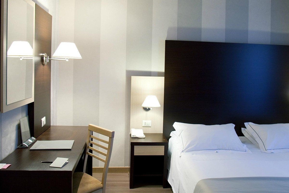 Een hotelkamer met een bed, een bureau, een stoel, een nachtkastje met lamp en een spiegel.