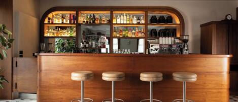Zona de bar con mostrador de madera, tres taburetes y estantes llenos de botellas y vasos.