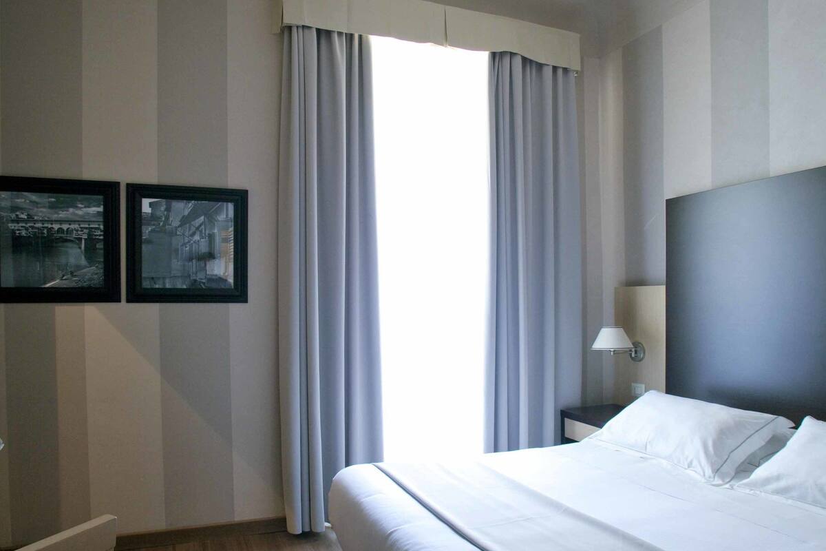 A hotel room with a large bed, a desk, two framed pictures on the wall, and a window with curtains.