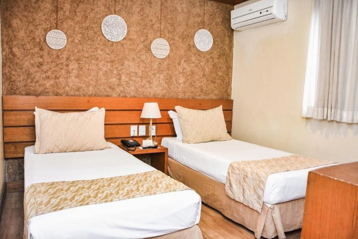A hotel room with two beds, a wooden headboard, a nightstand, a lamp, and a wall-mounted air conditioner.