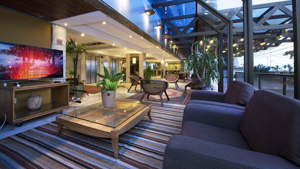 A modern hotel lobby with a large TV, comfortable seating, and a glass ceiling. - Magna Praia Hotel (Fortaleza)