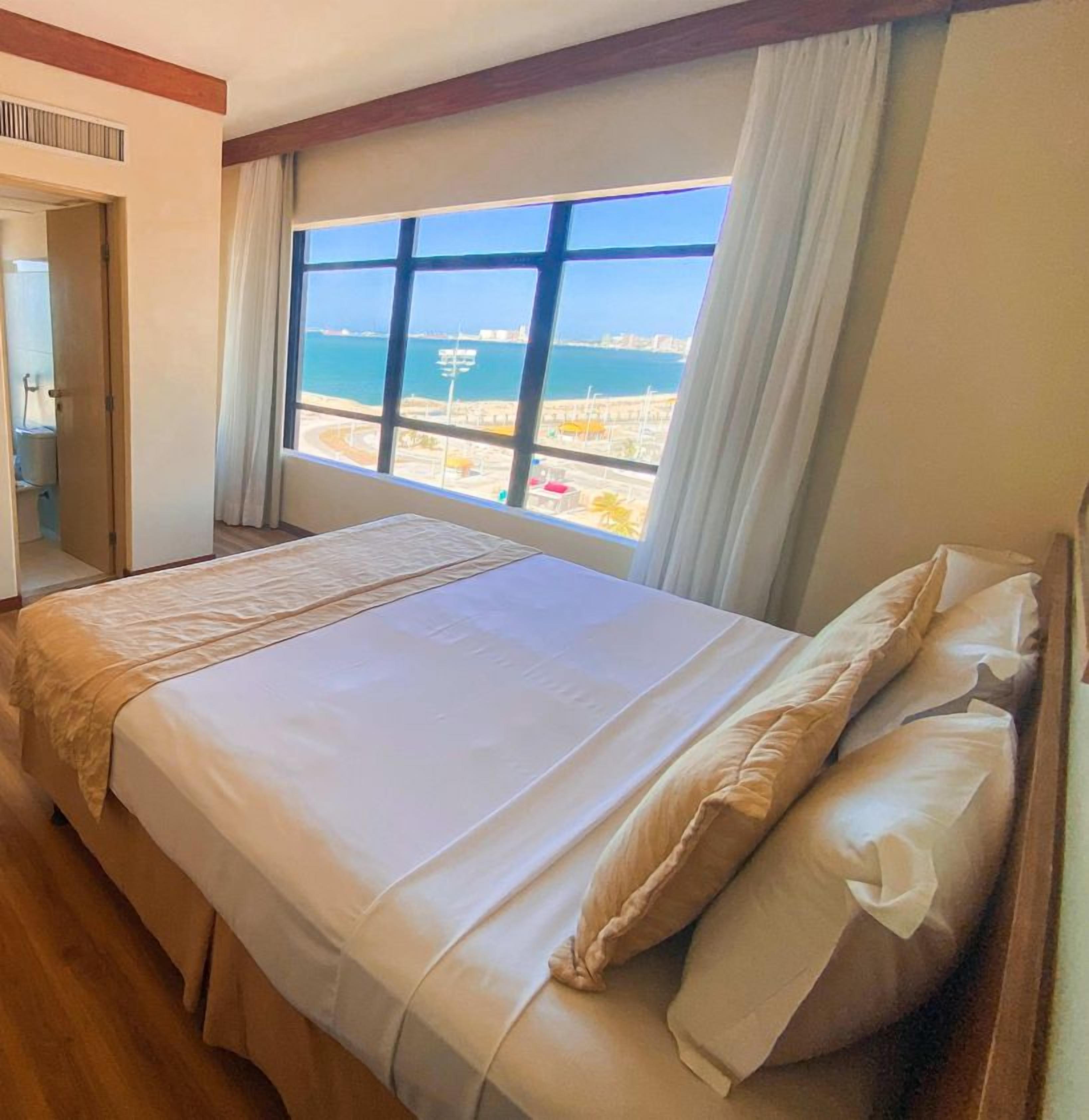 A hotel room with a large bed, a view of the sea, and a window with curtains.