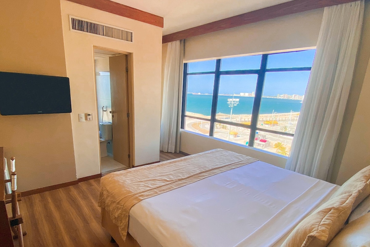A hotel room with a large bed, wooden flooring, a view of the sea, and a window with curtains.
