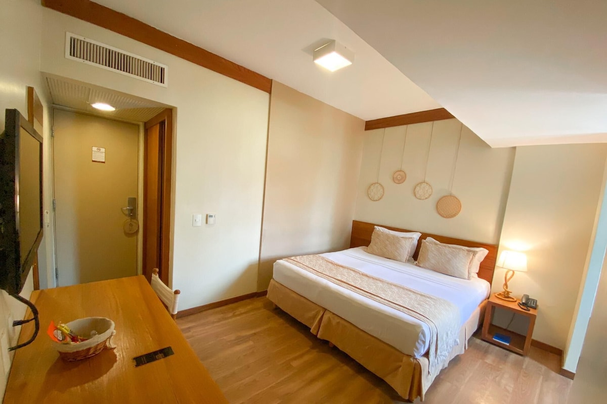 A hotel room with a bed, bedside table, lamp, and a wooden desk with a basket.