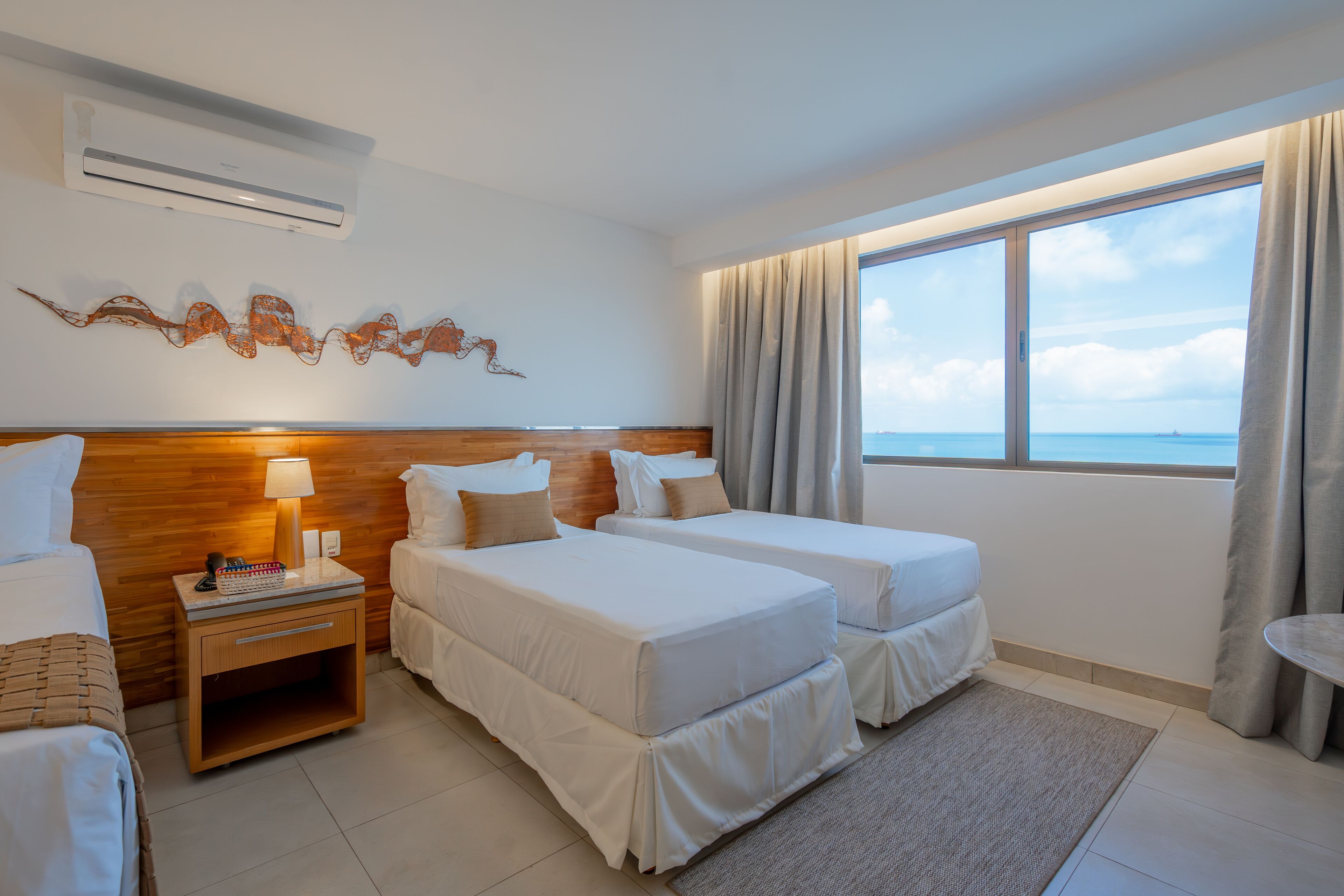 A hotel room with two beds, a wooden headboard, a nightstand with a lamp, and a view of the sea through large windows.