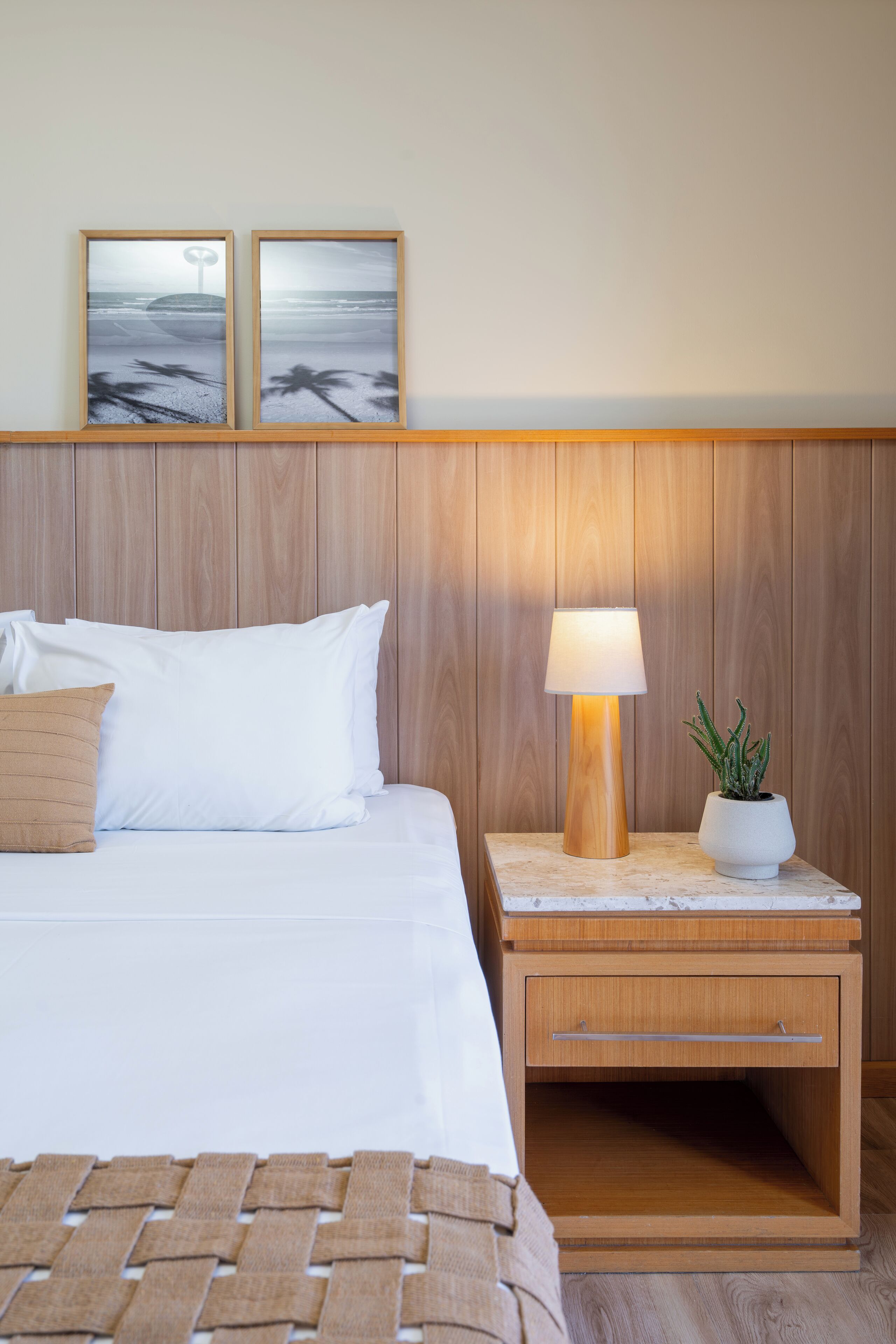 Un lit bien fait, avec du linge de lit blanc et un oreiller marron, une table de chevet avec une lampe et une plante en pot, et deux tableaux encadrés accrochés au mur.