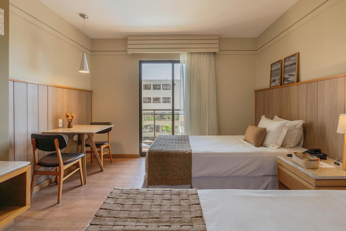 A hotel room with two beds, a desk, and a chair. There is a window with a view of buildings outside.