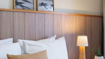 Habitación de hotel con una cama, almohadas blancas, un cojín color beige, un cabecero de madera, una mesita de noche con una lámpara y una planta en maceta.