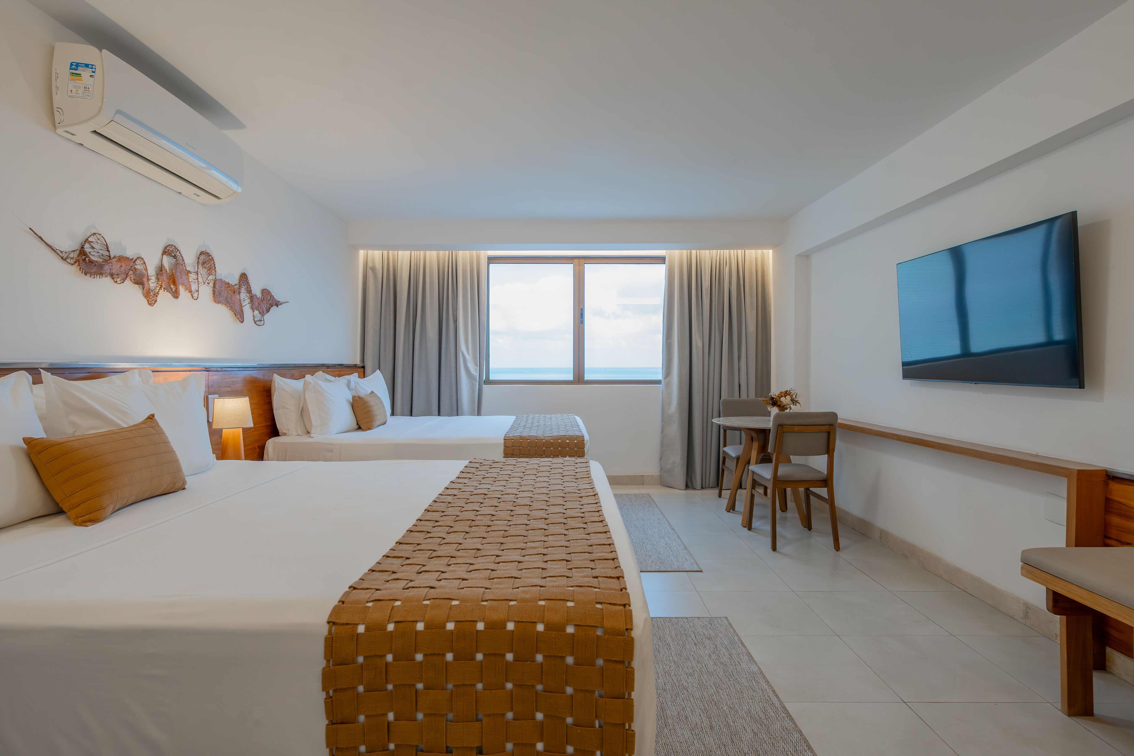 Quarto de hotel com duas camas, televisão, janela com cortinas e vista para o mar.