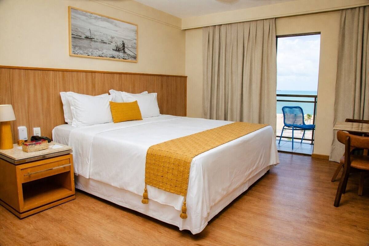 A hotel room with a large bed, a wooden nightstand, a desk, and a chair. There is a window with curtains and a view of the sea.