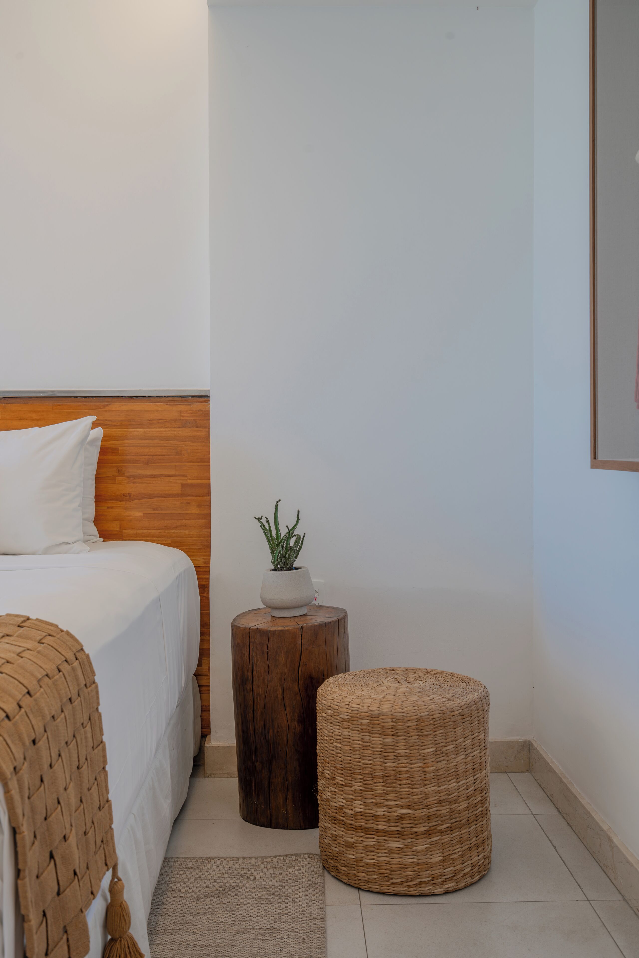 A bedroom with a wooden headboard, a bed with white bedding, a small round wooden table with a plant, and a wicker ottoman.