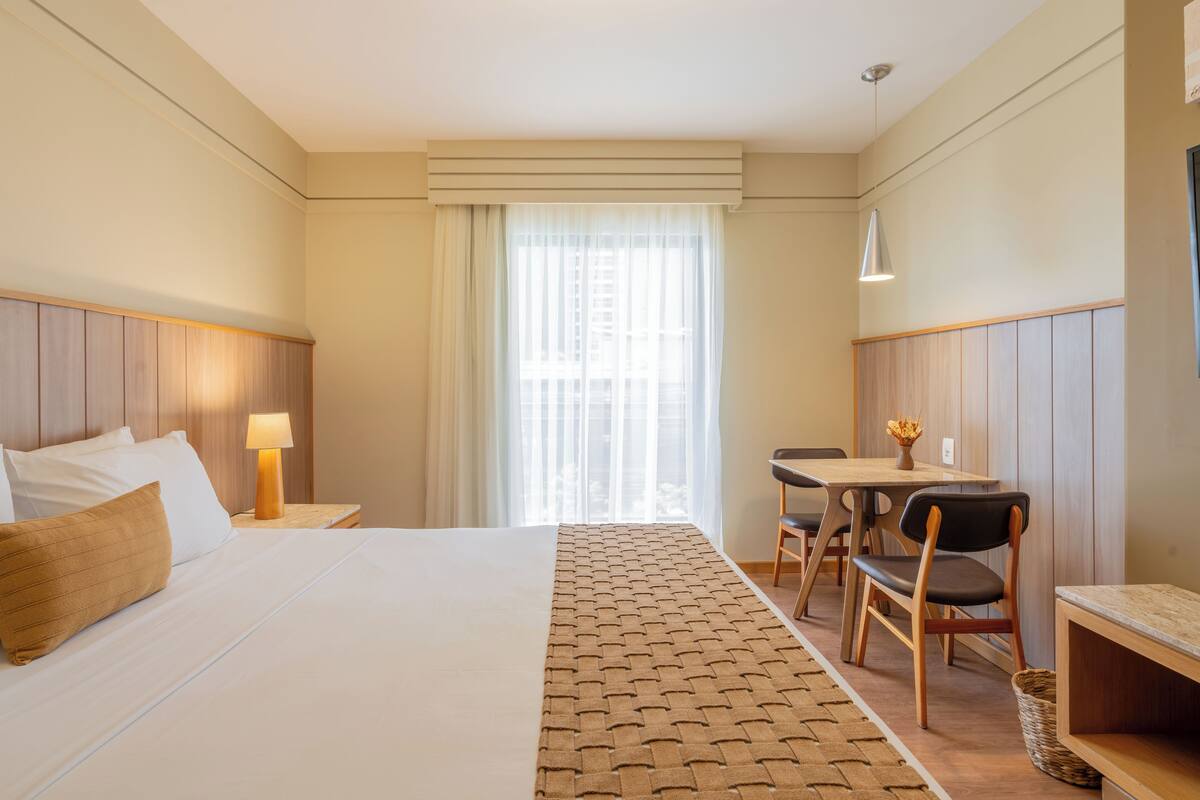 A hotel room with a large bed, a desk with a chair, a lamp, and a window with sheer curtains.