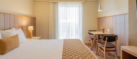 Quarto de hotel com uma cama grande, uma escrivaninha com cadeira, um abajur e uma janela com cortinas finas.