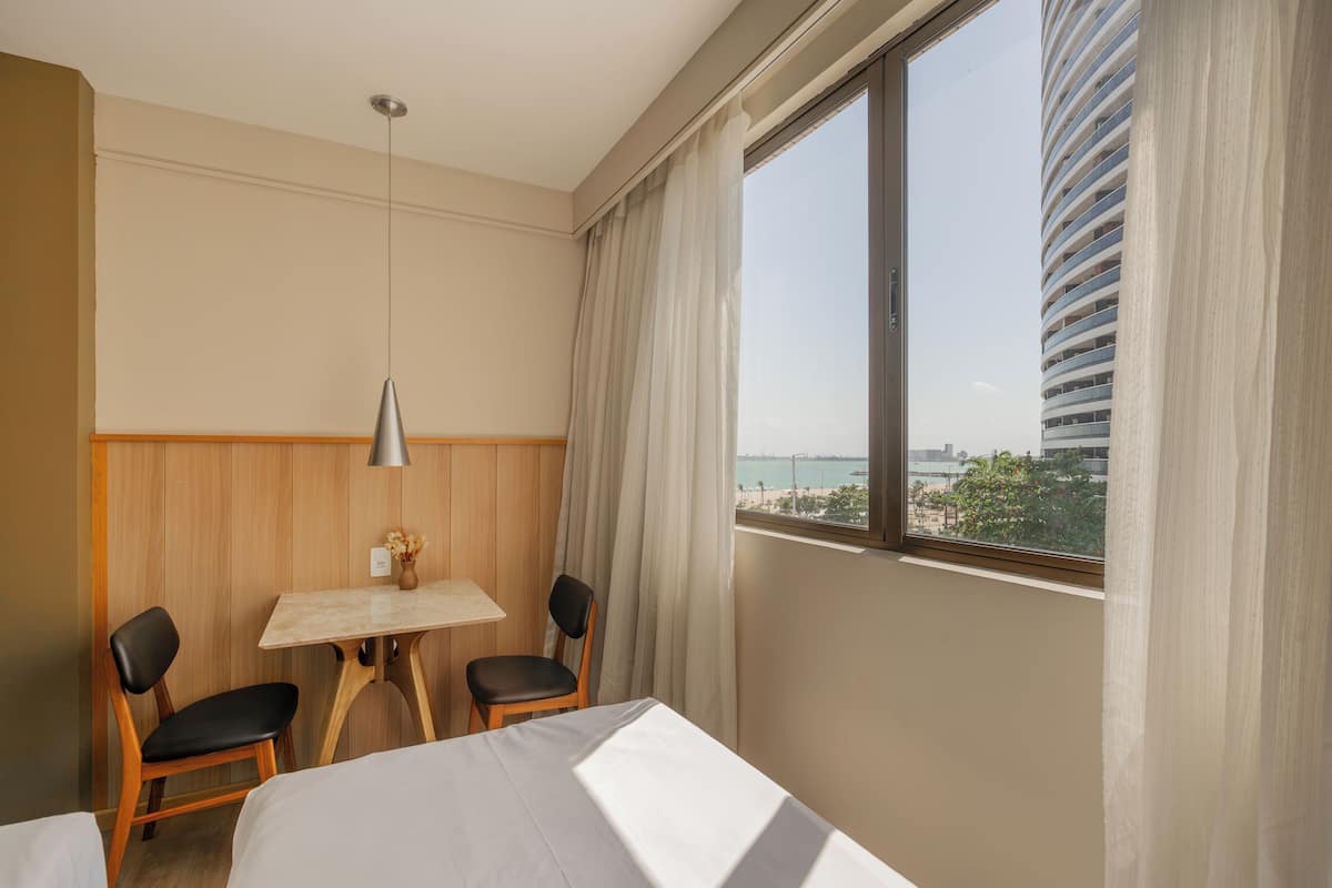 A hotel room with a bed, two chairs, a small table with a vase, a lamp, and a large window with a view of a building and the sea.