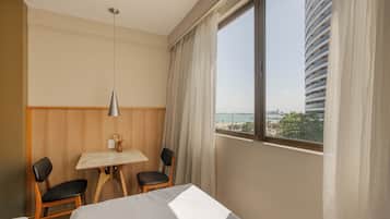 Ein Hotelzimmer mit einem Bett, zwei Stühlen, einem kleinen Tisch mit einer Vase, einer Lampe und einem großen Fenster mit Blick auf ein Gebäude und das Meer.