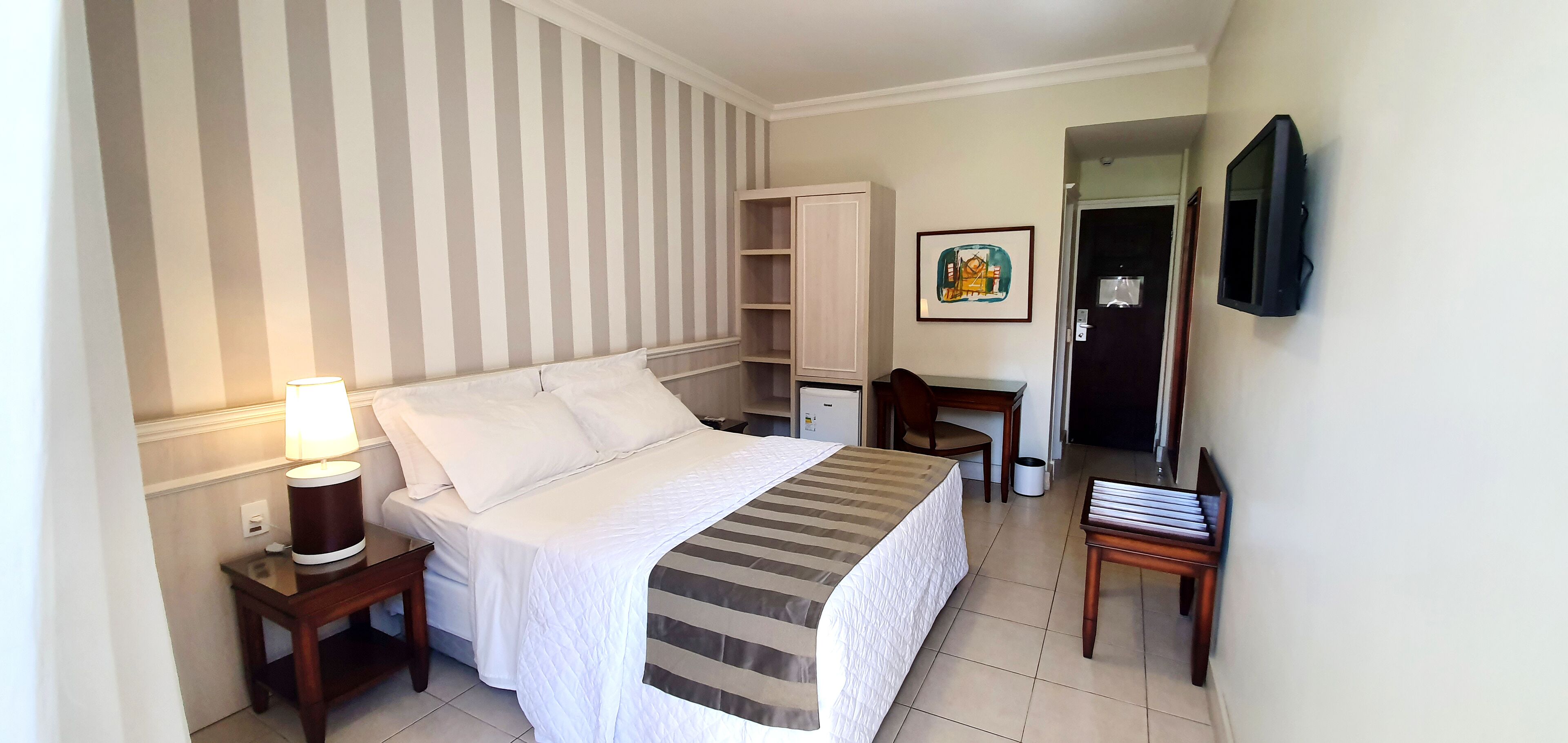 A hotel room with a bed, bedside table with a lamp, a desk with a chair, a TV mounted on the wall, and a door leading to another room.