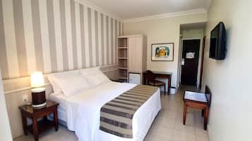 A hotel room with a bed, bedside table with a lamp, a desk with a chair, a TV mounted on the wall, and a door leading to another room.