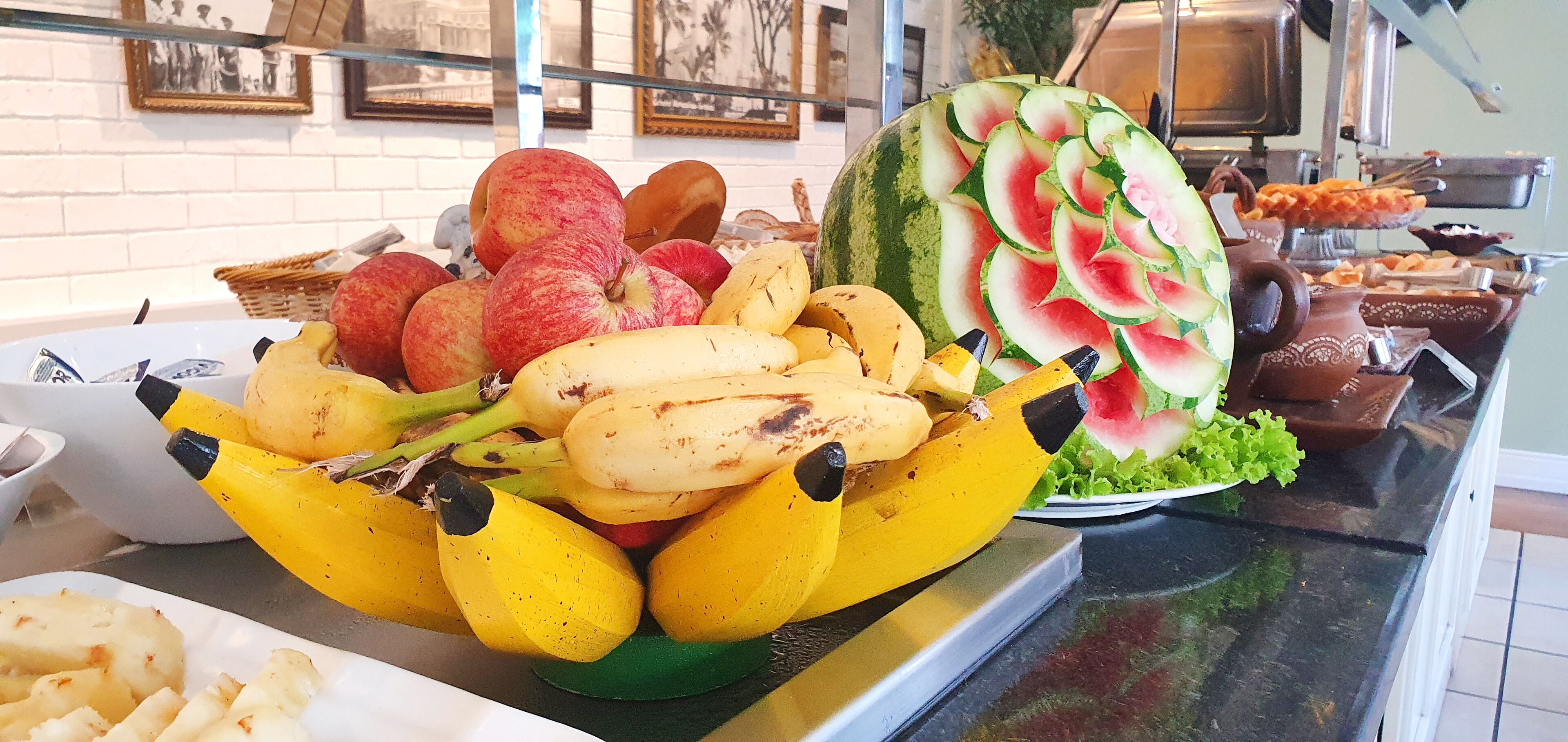 Una exhibición de frutas con plátanos, manzanas y una sandía tallada con forma de flor.