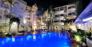 A swimming pool with lounge chairs and umbrellas, surrounded by palm trees and buildings with balconies.