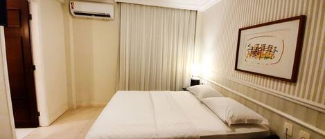 A hotel room with a bed, a nightstand, a lamp, a framed picture on the wall, and an air conditioning unit.
