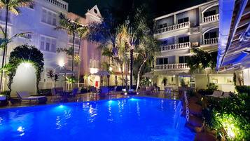 Uma área de piscina à noite com espreguiçadeiras e guarda-sóis, cercada por palmeiras e um edifício com varandas.