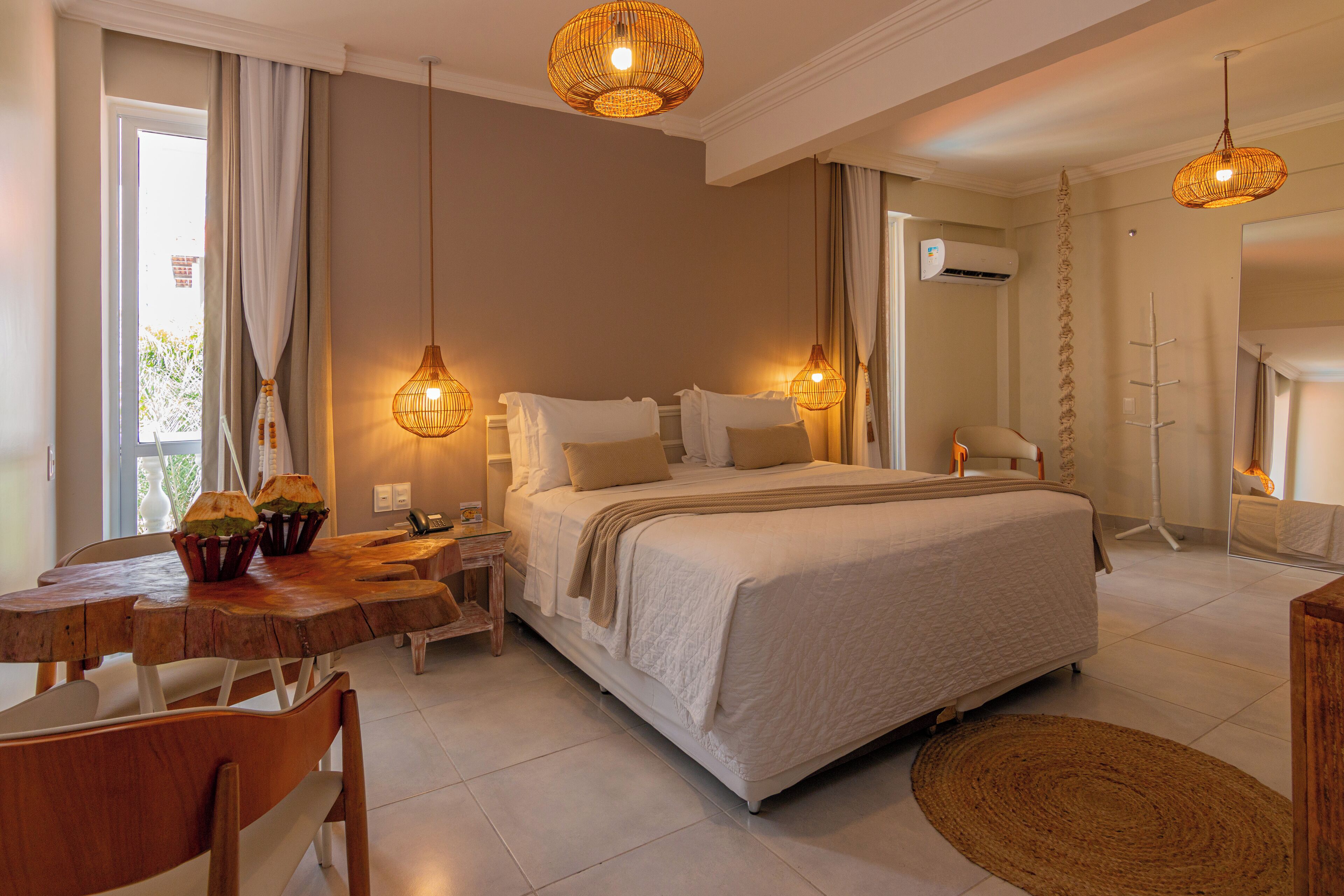 A bedroom with a large bed, two bedside tables, a wooden dining table, and a chair. There are two hanging lamps, a window with curtains, and a mirror on the wall.