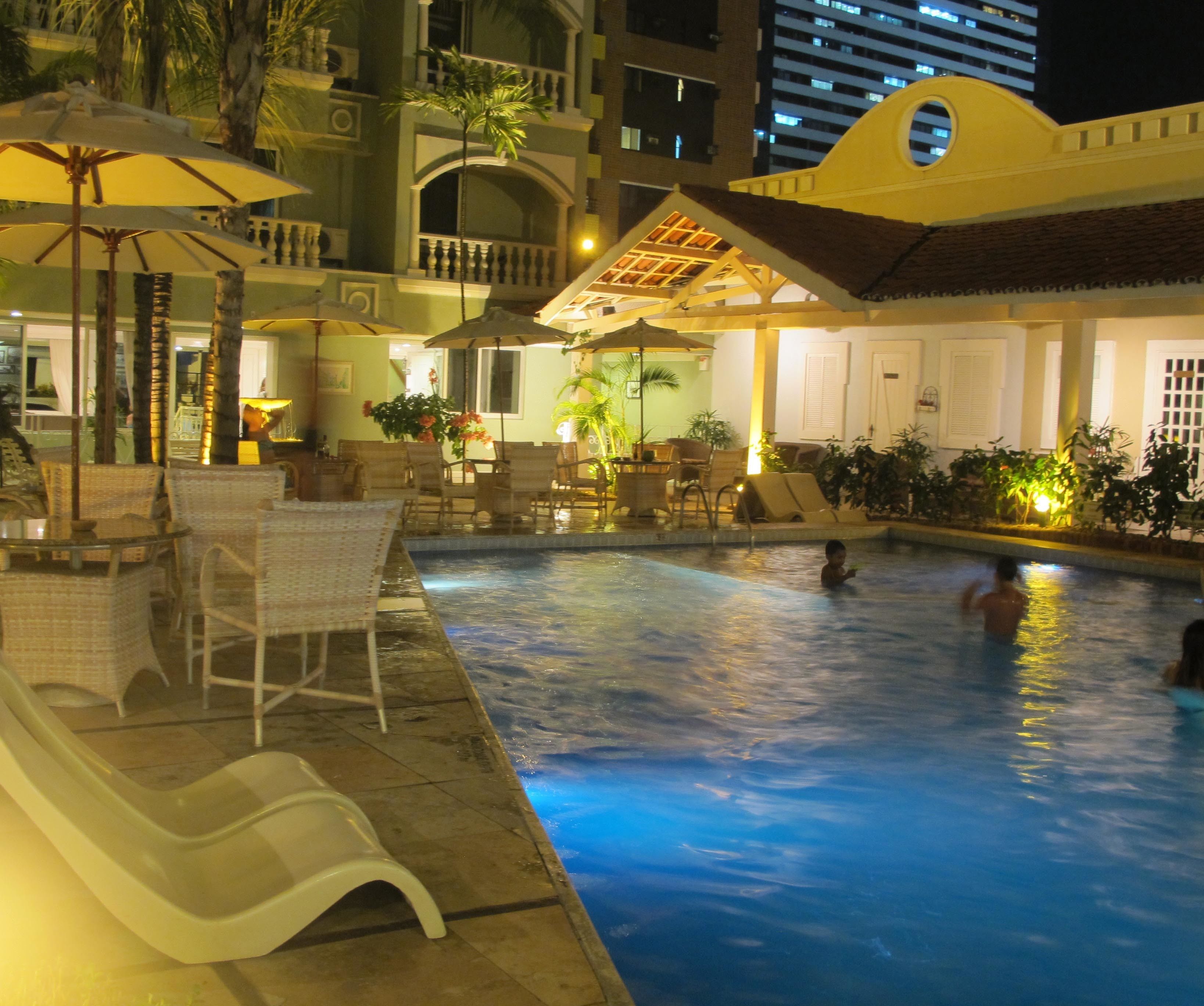 A swimming pool area with lounge chairs and umbrellas, surrounded by a building with balconies and windows.