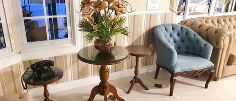 A room with a tufted blue armchair, a glass table with a vase of dried flowers, and a vintage telephone.