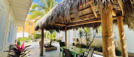 A covered outdoor area with a table and chairs, surrounded by plants and a thatched roof structure.