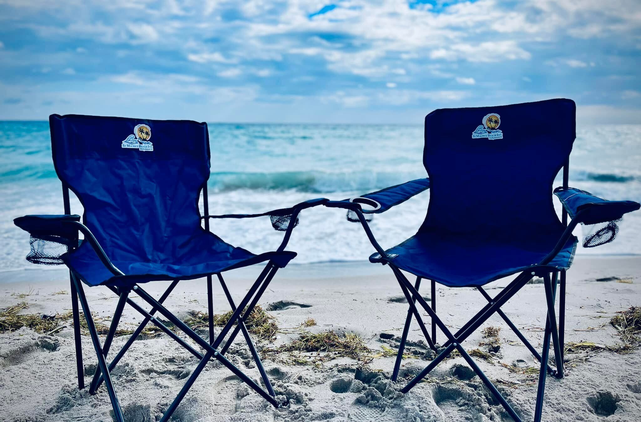 Zwei blaue Klappstühle mit Logo an der Rückenlehne, auf einem sandigen Strand mit Blick aufs Meer.