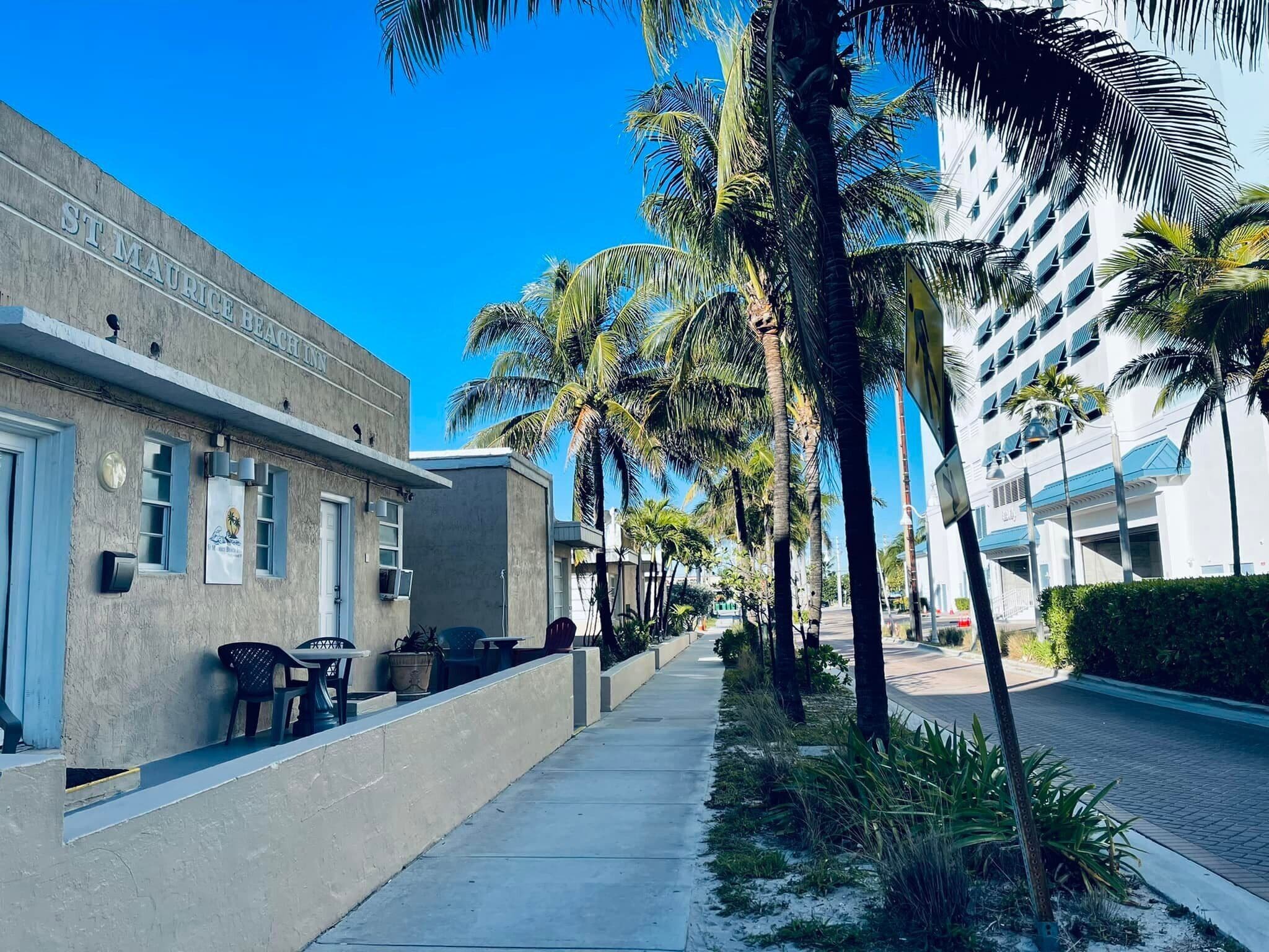 A hotel named St. Maurice Beach with a concrete facade, outdoor seating, and palm trees lining the street.
