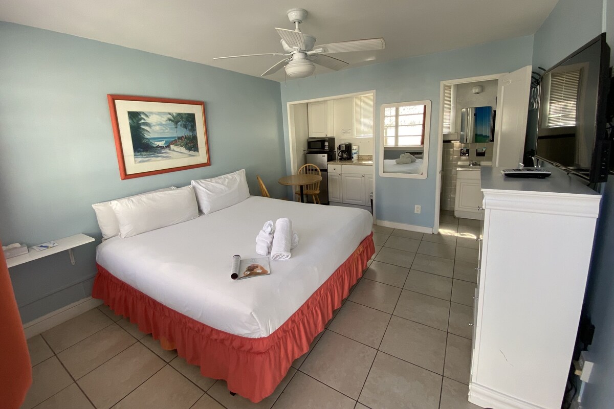 A hotel room with a bed, a ceiling fan, a painting, a kitchenette, and a bathroom visible through an open door.