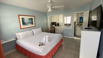 Habitación de hotel con cama, ventilador de techo, un cuadro, kitchenette y baño visible a través de una puerta abierta.